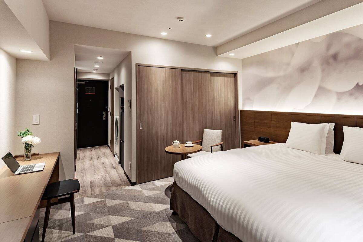 A hotel room with a bed, a desk with a laptop, a chair, a small table with a teapot, and a wardrobe.
