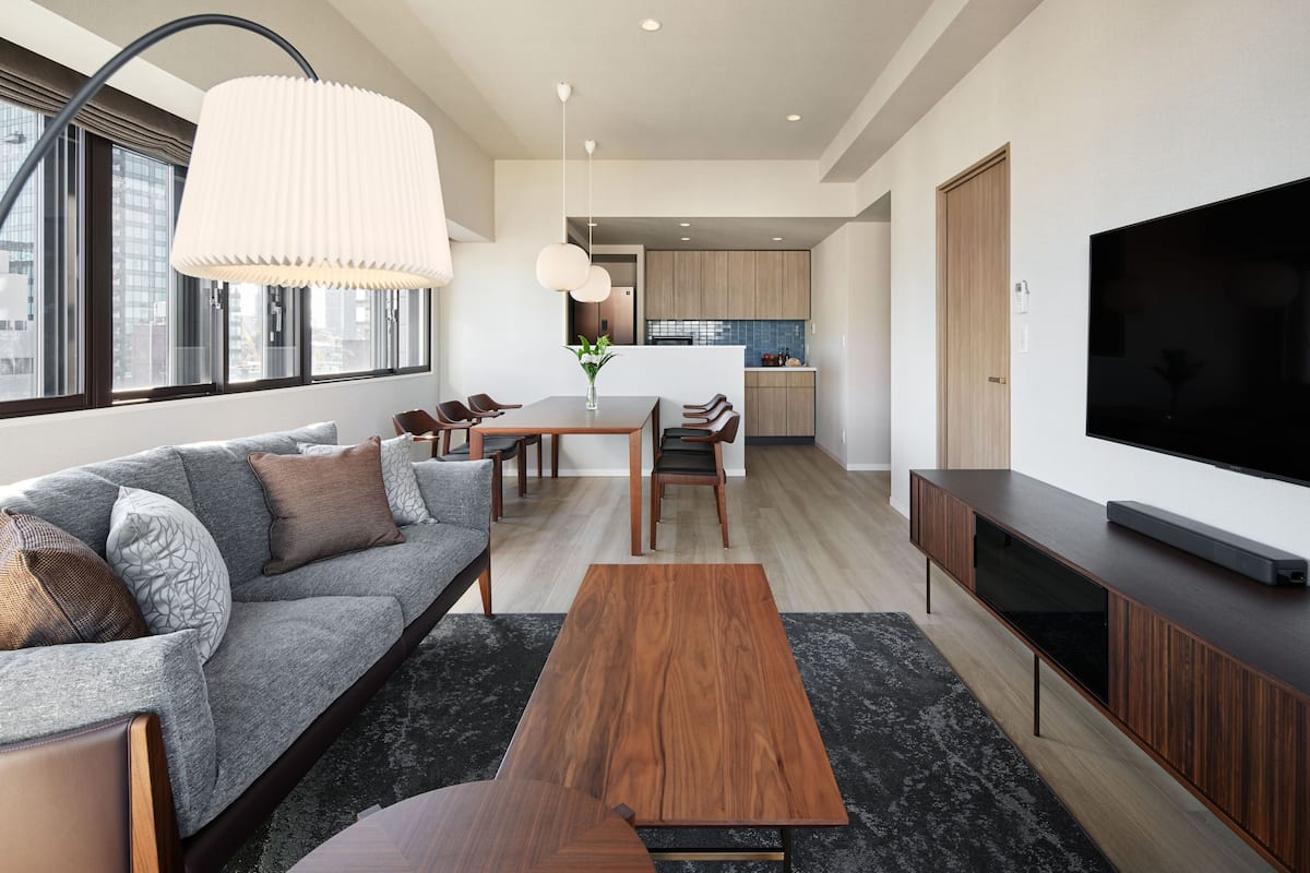 A modern living room with a sofa, coffee table, and TV stand. There is a dining area with chairs and a table, and a kitchen in the background.