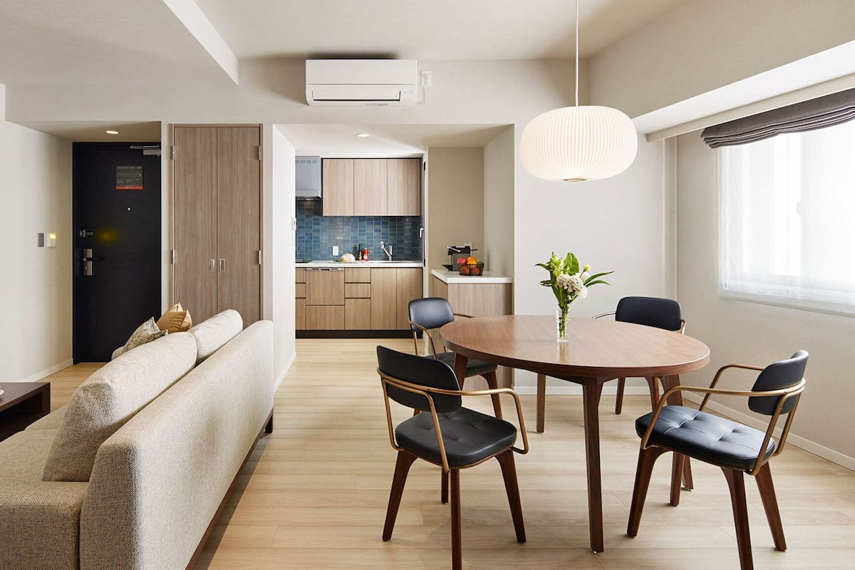 A modern living room with a sofa, dining table, and chairs. There is a kitchen in the background with wooden cabinets and a tiled backsplash.