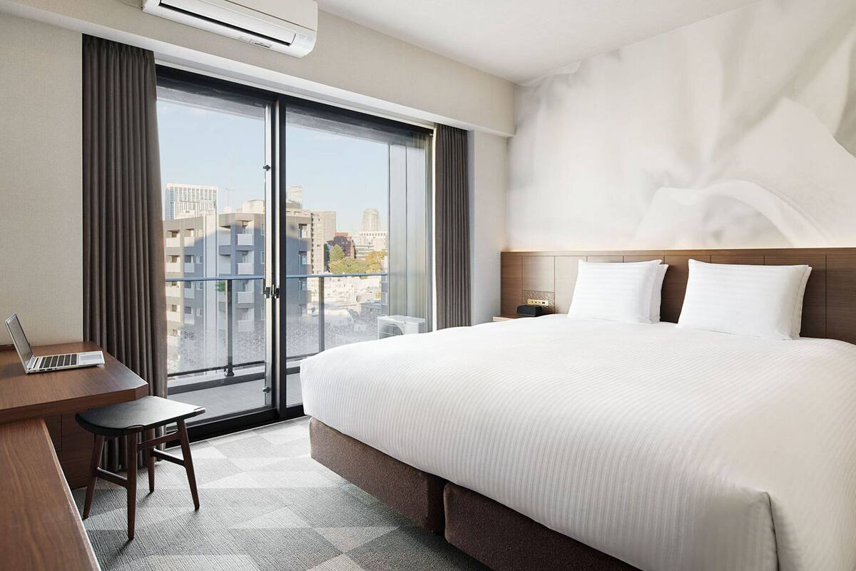 A hotel room with a large bed, a desk, and a view of the city through the window.