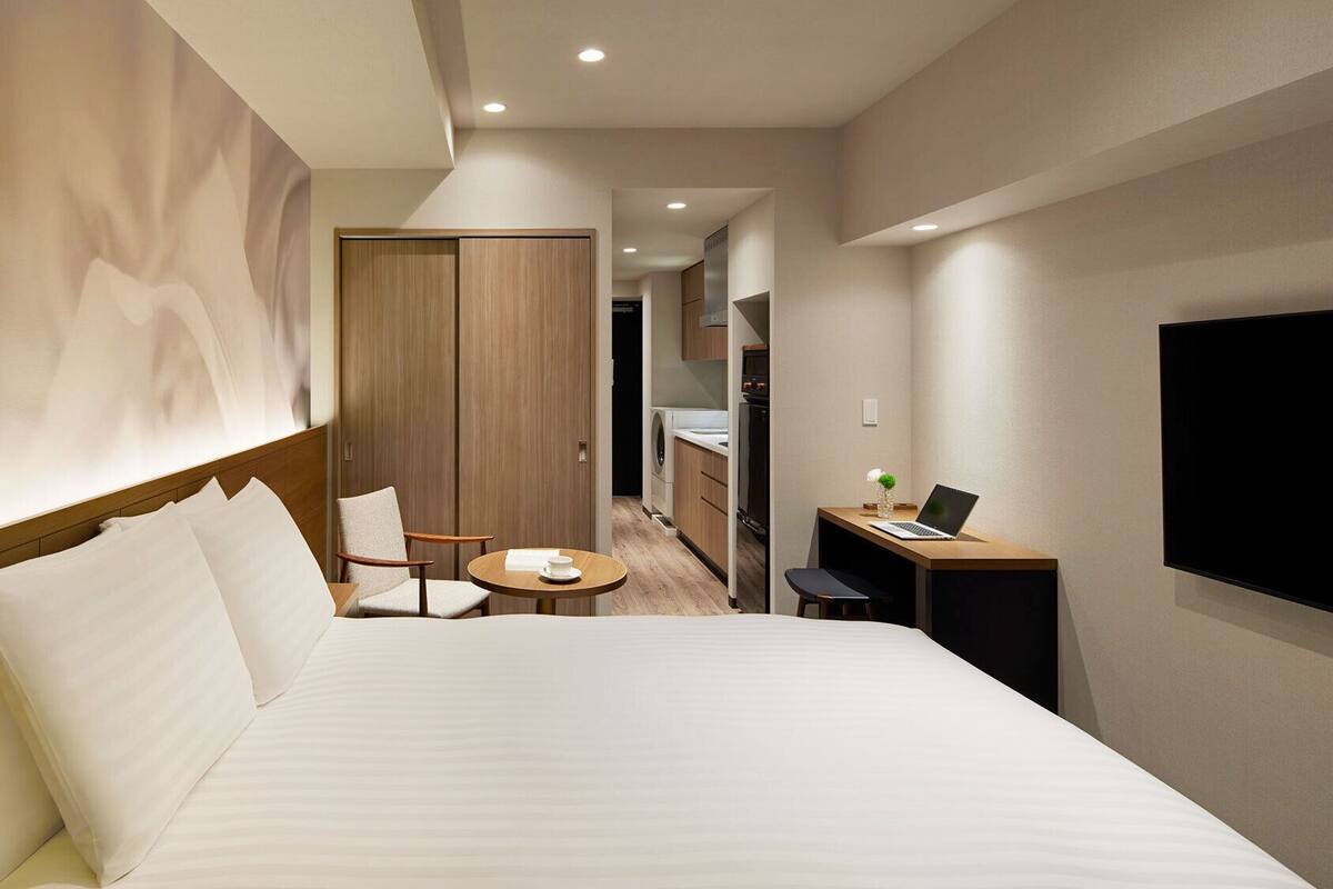 A hotel room with a large bed, a desk with a laptop, a TV, and a small dining table.