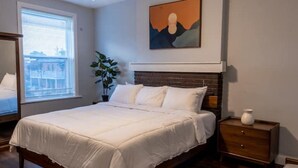 A bedroom with a large bed, a nightstand, a potted plant, and a painting on the wall.
