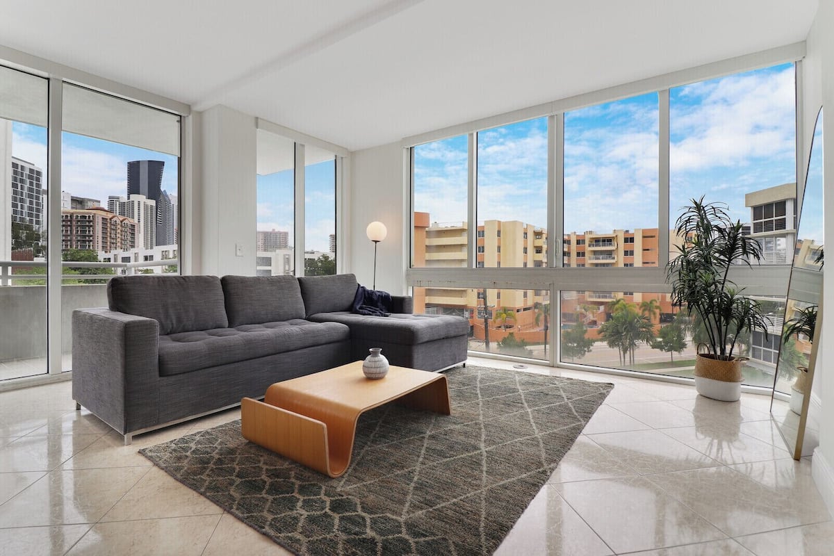 A modern living room with a grey sectional sofa, a wooden coffee table, and a large window offering a city view.