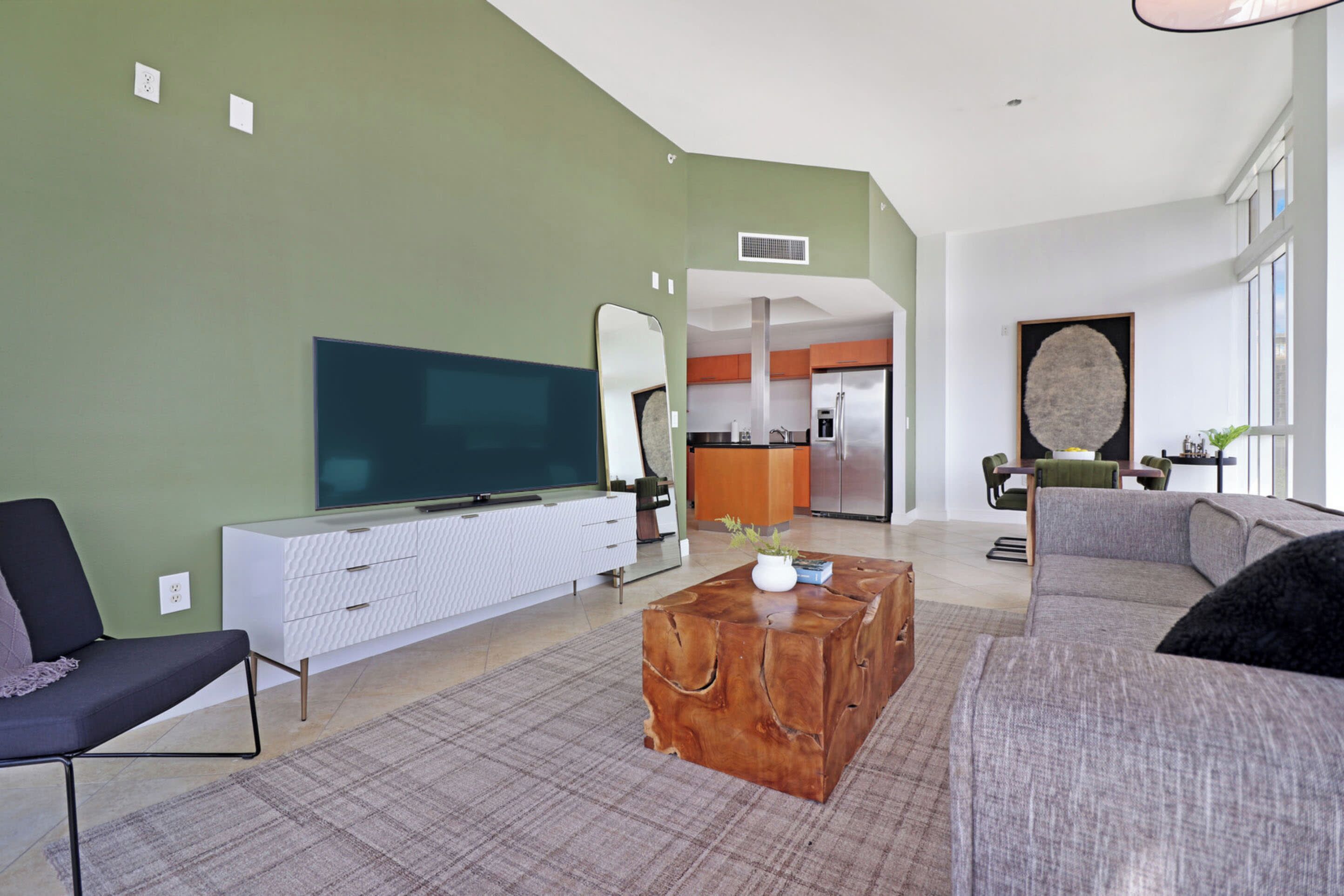 A modern living room with a flat-screen TV, a wooden coffee table, a grey sofa, and a dining area in the background.
