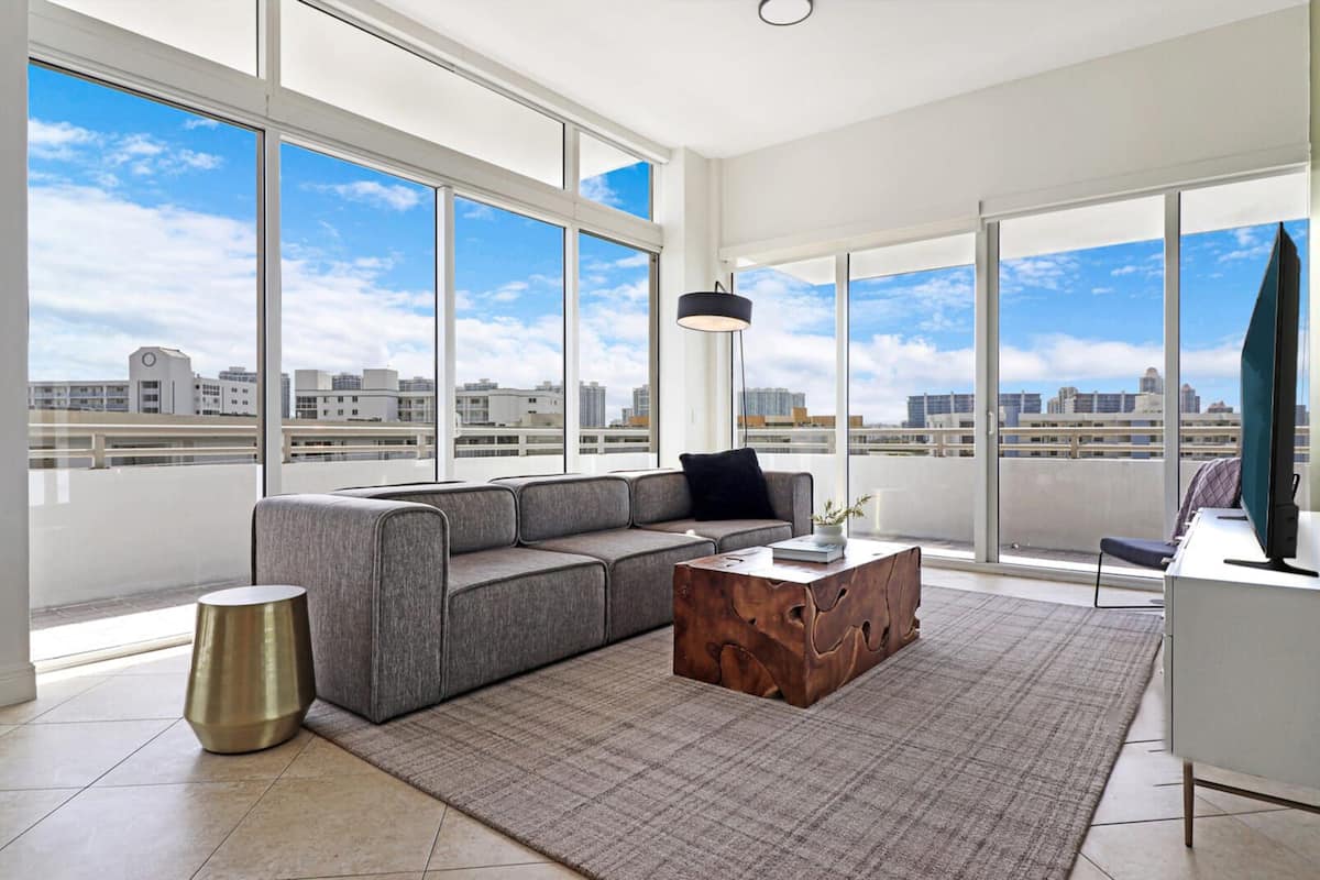 A modern living room with a grey sectional sofa, a wooden coffee table, and a view of the cityscape through large windows.
