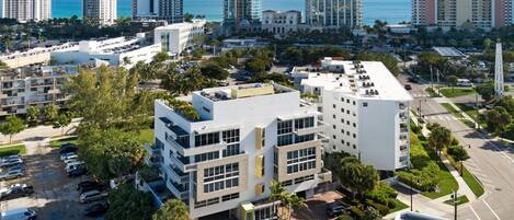 A modern building with a flat roof, surrounded by parking and greenery, near high-rise buildings and a coastline.