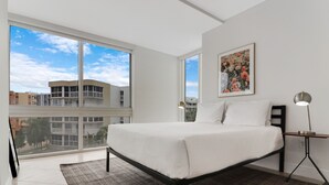 Une chambre moderne avec un grand lit, une vue sur les immeubles à travers de grandes fenêtres du sol au plafond et un tableau encadré accroché au mur.