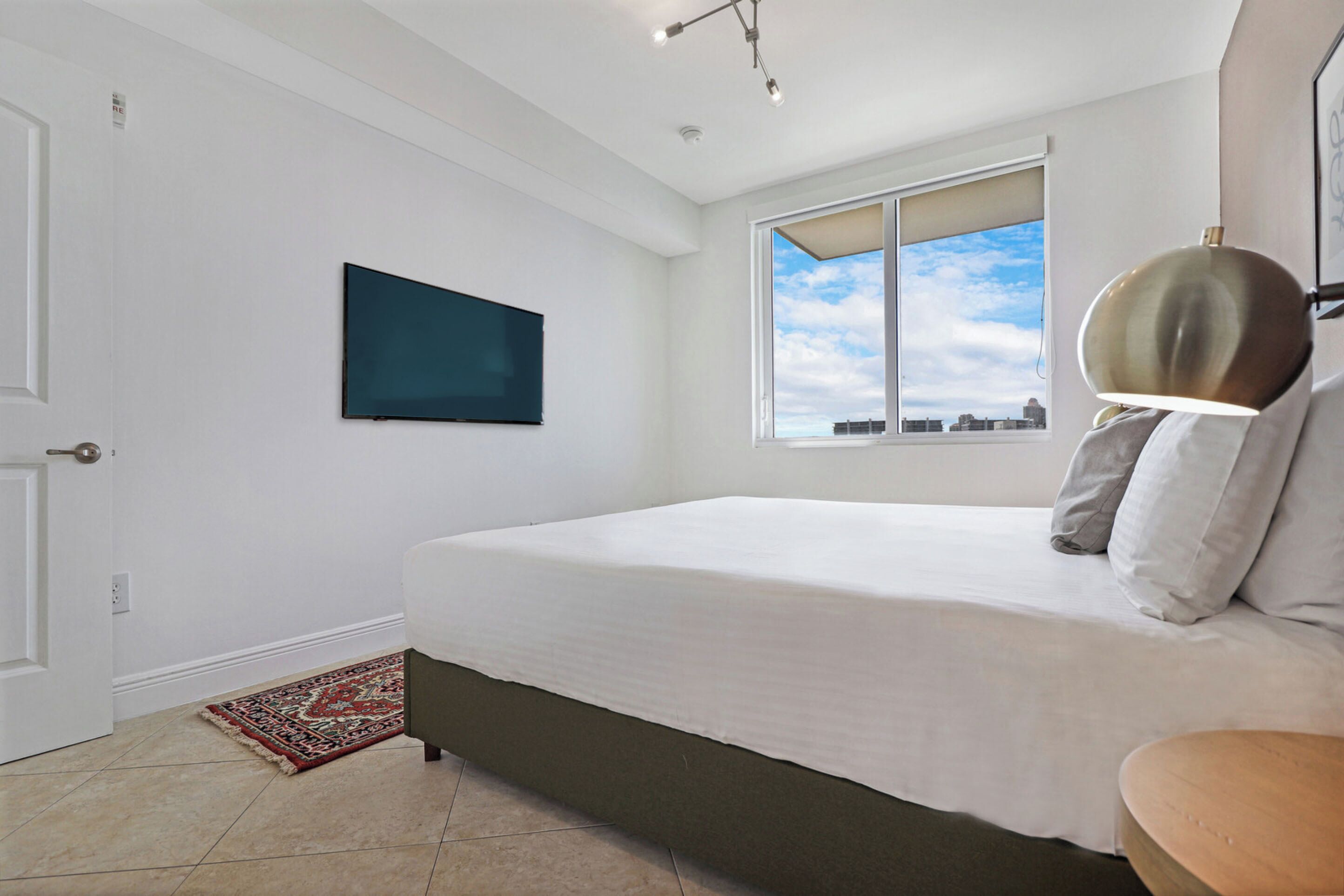 A bedroom with a bed, a window with a city view, a mounted TV, and a door.