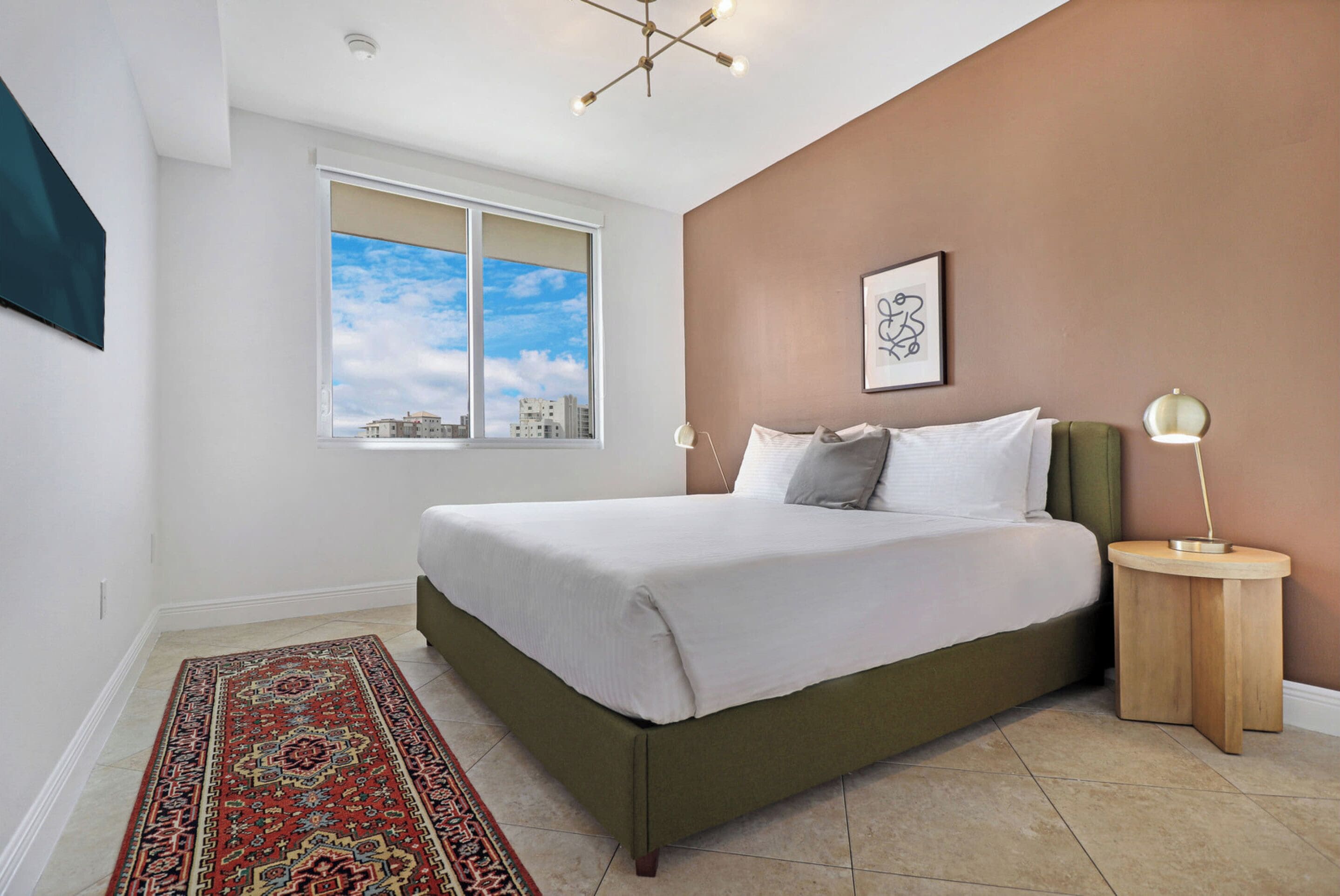Un dormitorio con una cama grande, una ventana con vista a la ciudad, una mesita de noche con una lámpara y una alfombra estampada.