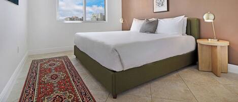Un dormitorio con una cama grande, una ventana con vista a la ciudad, una mesita de noche con una lámpara y una alfombra estampada.