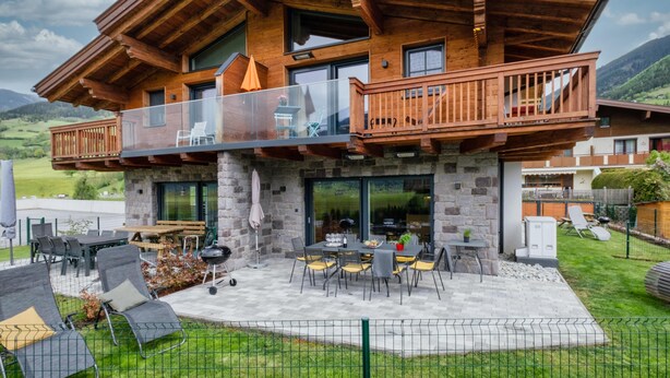 A two-story wooden chalet with a balcony, outdoor seating, and a stone foundation.