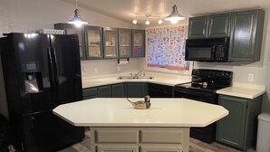 A kitchen with a black refrigerator, a microwave, a stove, and a dishwasher. There is a large island with a sink and a window above it. The cabinets are green, and the countertops are white. - Cabin in the Pines - 3 Minutes to the beach, docks or golf! (Cascade)