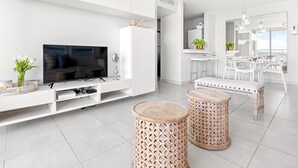Een moderne woonkamer met een flatscreen televisie, witte kasten, een eethoek met een witte tafel en stoelen, en twee decoratieve krukken.