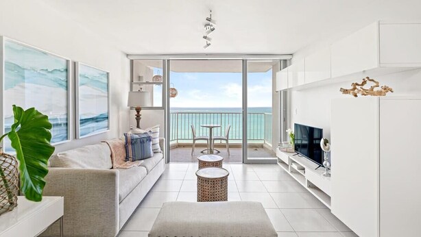 Een moderne woonkamer met een bank, salontafel en televisie, die uitkomt op een balkon met uitzicht op de oceaan.