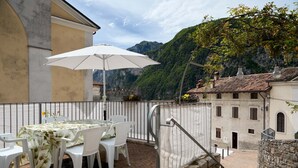 Eine Terrasse im Freien mit Tisch und Stühlen, einem weißen Sonnenschirm und Blick auf Berge und Gebäude.