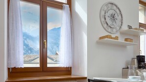 Ein Zimmer mit Holzfenster, weißen Vorhängen, einer Wanduhr, einem Tisch mit Kaffeemaschine und Blick auf die Berge.
