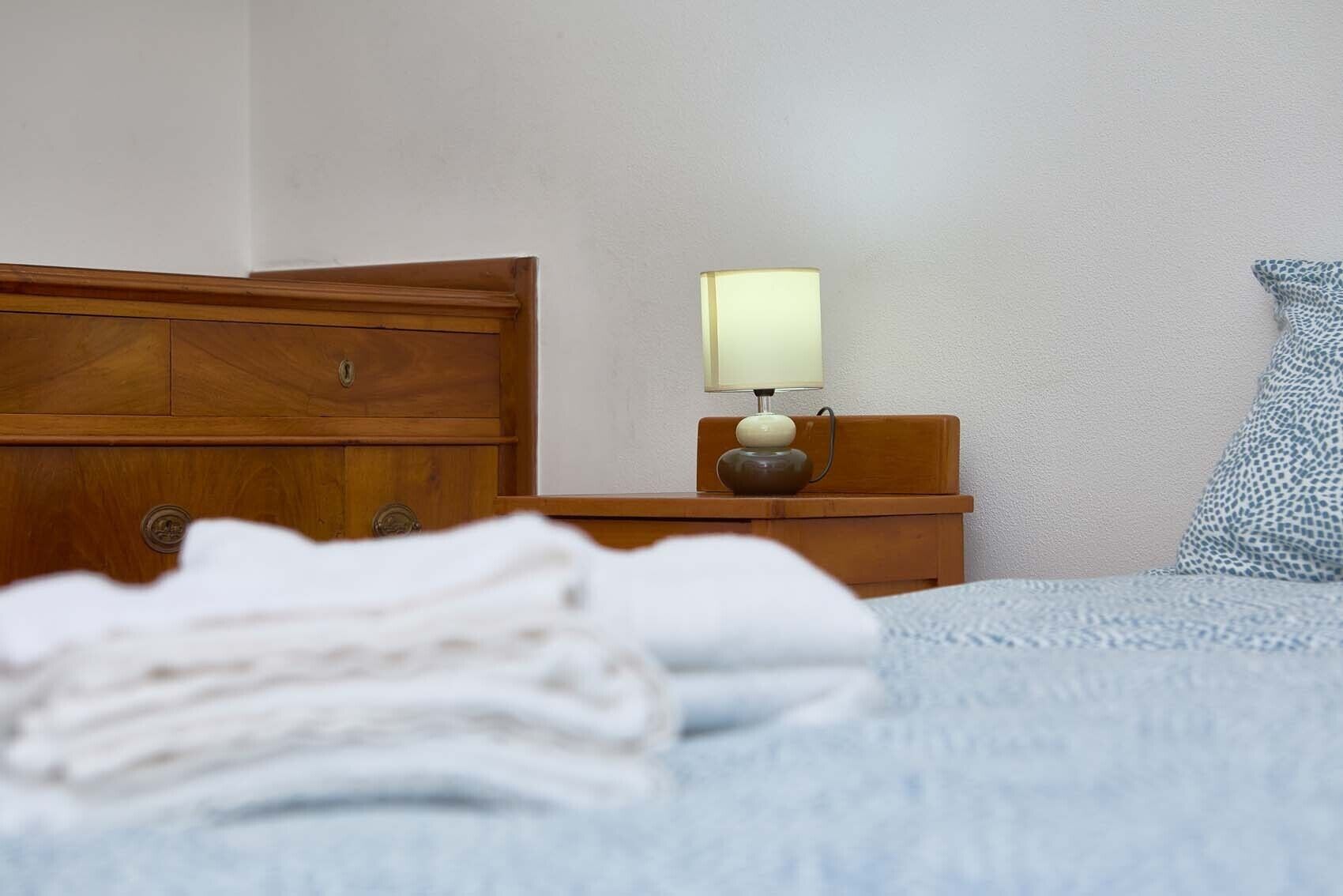 Ein ordentlich bezogenes Bett mit einem gefalteten weißen Handtuch, ein Nachttisch aus Holz mit einer Lampe und ein gemustertes Kopfkissen.