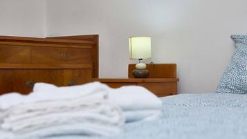 Ein ordentlich bezogenes Bett mit einem gefalteten weißen Handtuch, ein Nachttisch aus Holz mit einer Lampe und ein gemustertes Kopfkissen.