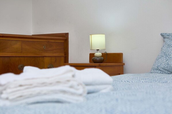A neatly made bed with a folded white towel, a wooden nightstand with a lamp, and a patterned pillow.