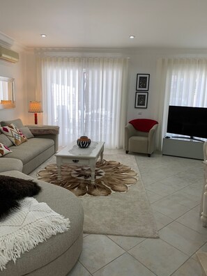 Una sala de estar con sofá, una mesa de centro, televisor y un ventanal con cortinas translúcidas.