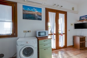 Une pièce équipée d’un lave-linge, d’un four à micro-ondes, d’un téléviseur posé sur un meuble, d’une porte avec des rideaux et d’un tableau représentant un paysage côtier.