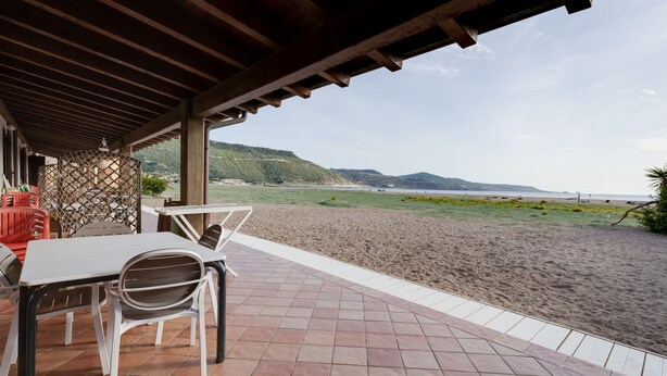 A covered patio with a table and chairs, overlooking a beach with a view of the sea and distant hills. - Holiday Home ‘Residenza Solemar - App 14’ with Sea View, Wi-Fi (Bosa Marina)