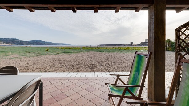 Vue sur la plage depuis un patio couvert, avec une table et des chaises, une pergola en bois et, au loin, une plage de sable.
