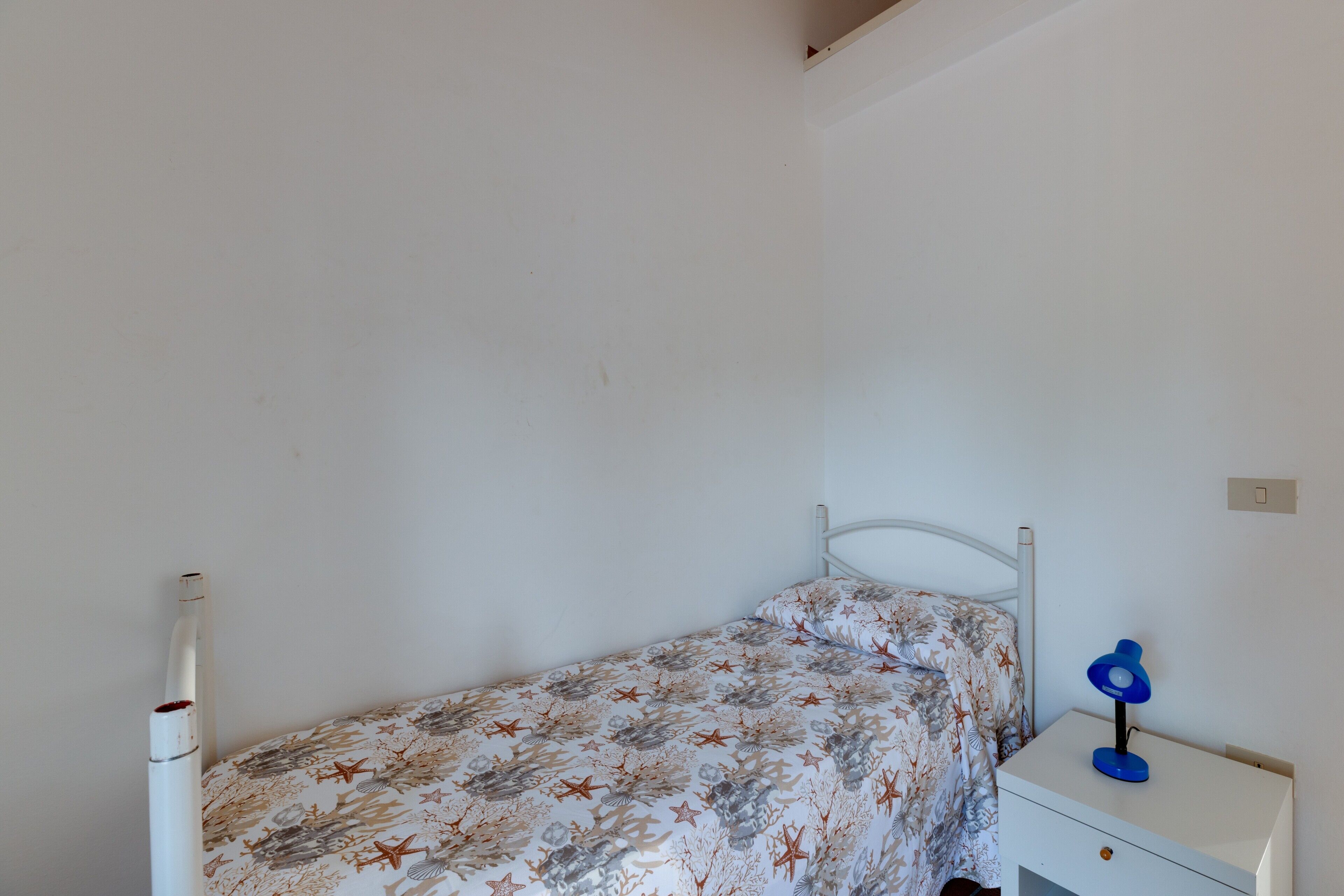 Una habitación pequeña con una cama individual, una colcha de motivos florales, una mesita de noche blanca con una lámpara azul y una pared blanca.