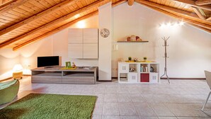 Una habitación en el ático con techo de madera inclinado, un televisor de pantalla plana, un escritorio, una estantería y un perchero.