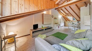 Una habitación moderna estilo loft con techo inclinado, un sofá gris, una pequeña mesa de comedor y una zona de cocina.