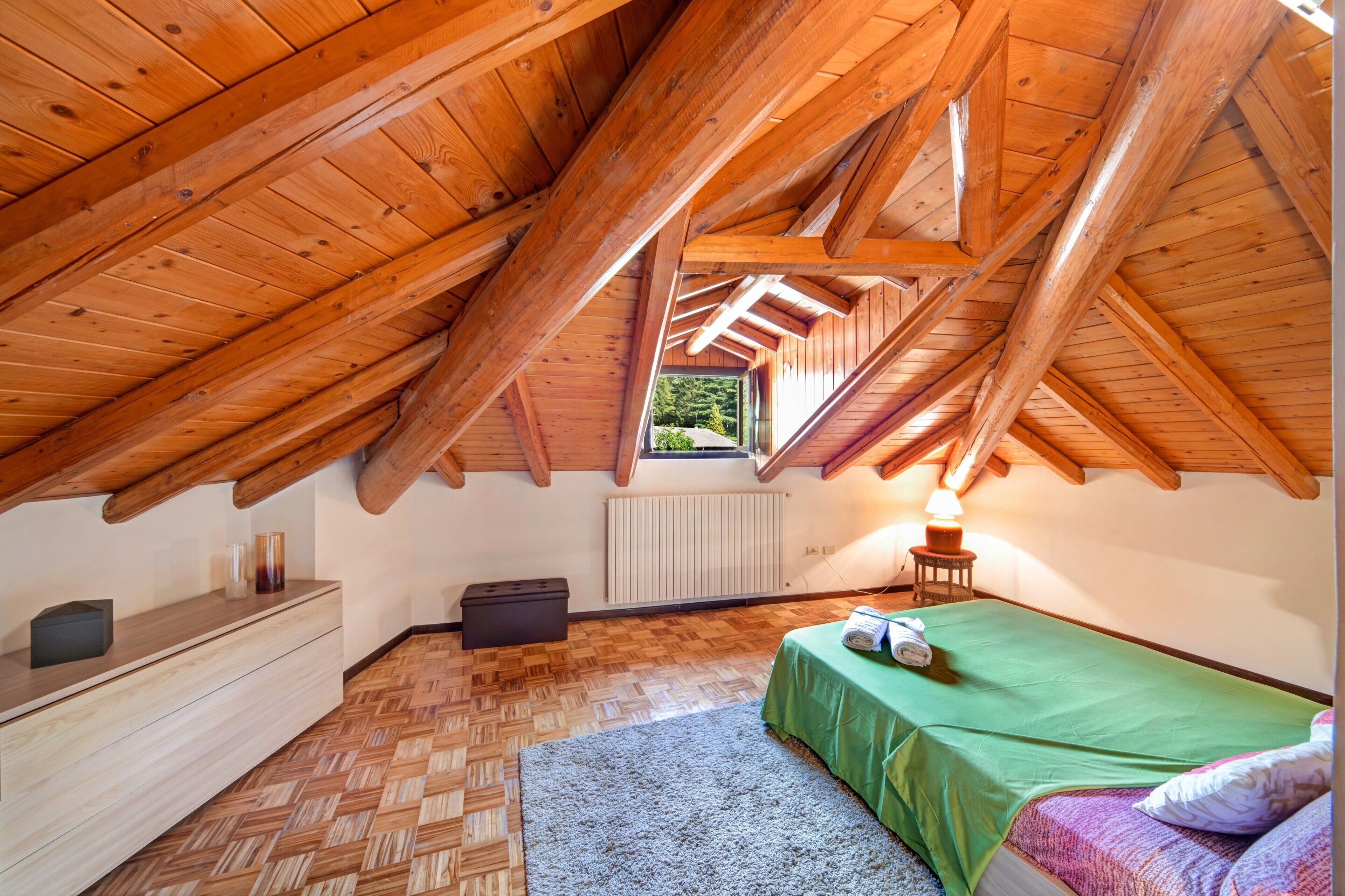 A room with a sloped wooden ceiling, a bed with a green bedspread, a small bedside table with a lamp, a radiator, and a herringbone-patterned floor.