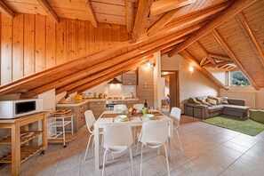 A modern loft with exposed wooden beams, a kitchen with white appliances, a dining area with white chairs, and a living space with a sofa.