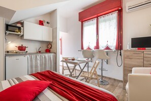 A compact room with a kitchenette, a dining area with a table and chairs, and a bed with red bedding.