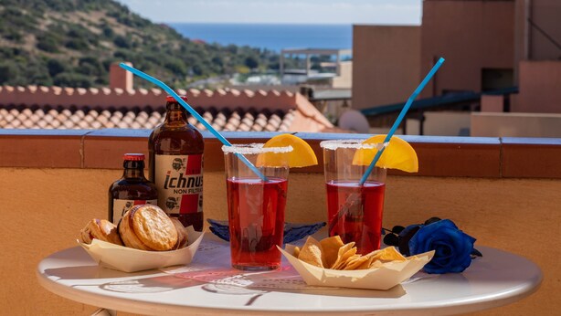 A table with drinks, snacks, and a bottle on a terrace with a view of the sea.