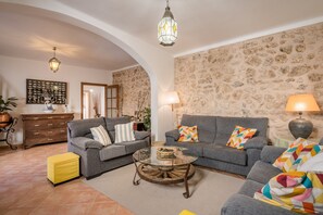Un salon avec deux canapés gris, une table basse en verre, un pouf jaune, un mur en pierre et un lustre.