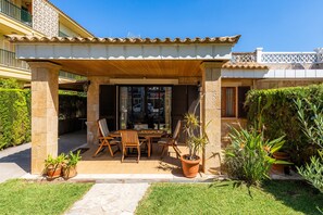 Un patio techado al aire libre con una mesa y sillas, rodeado de plantas en macetas y un muro de piedra.