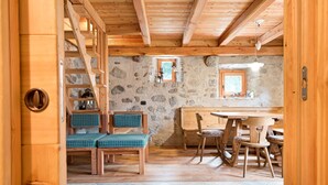 Een houten interieur met een stenen muur, een houten trap, een eettafel met stoelen en een bank met groene kussens.