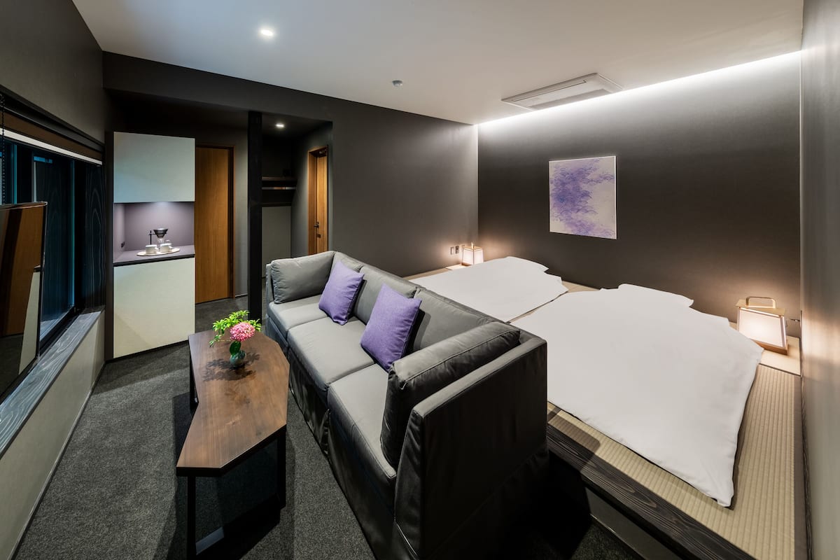 Een moderne hotelkamer met een slaapbank, een klein eettafeltje met een bloemenvaas, een nachtkastje met een lamp en een schilderij aan de muur.