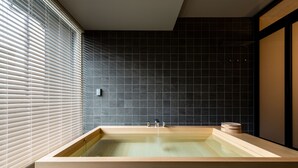 Een moderne badkamer met een houten badkuip, donkere betegelde muren en een raam met zonwering.