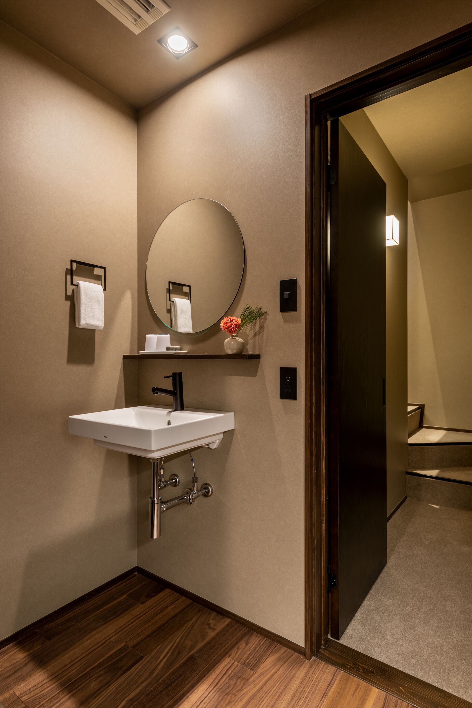 A modern bathroom with a white sink, a round mirror, and a wooden floor.