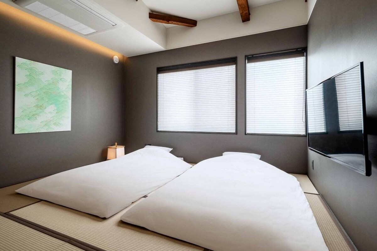 A room with a bed, a wall-mounted TV, and two windows with blinds.