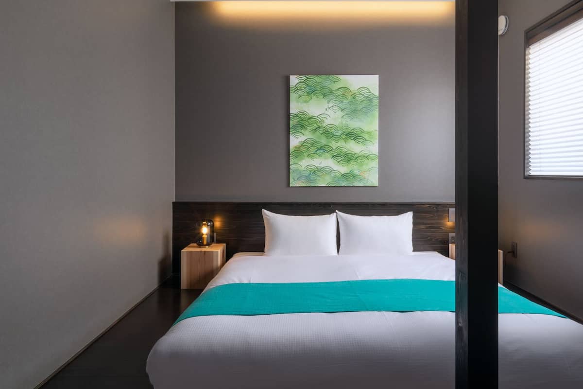 Een moderne slaapkamer met een groot bed, een plafond met houten balken, een groen abstract schilderij en een raam met zonwering.