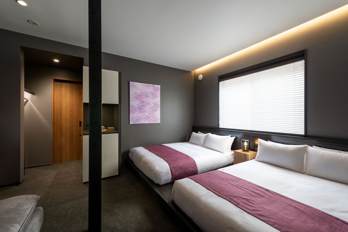 A modern hotel room with two beds, a desk, and a large window with blinds.