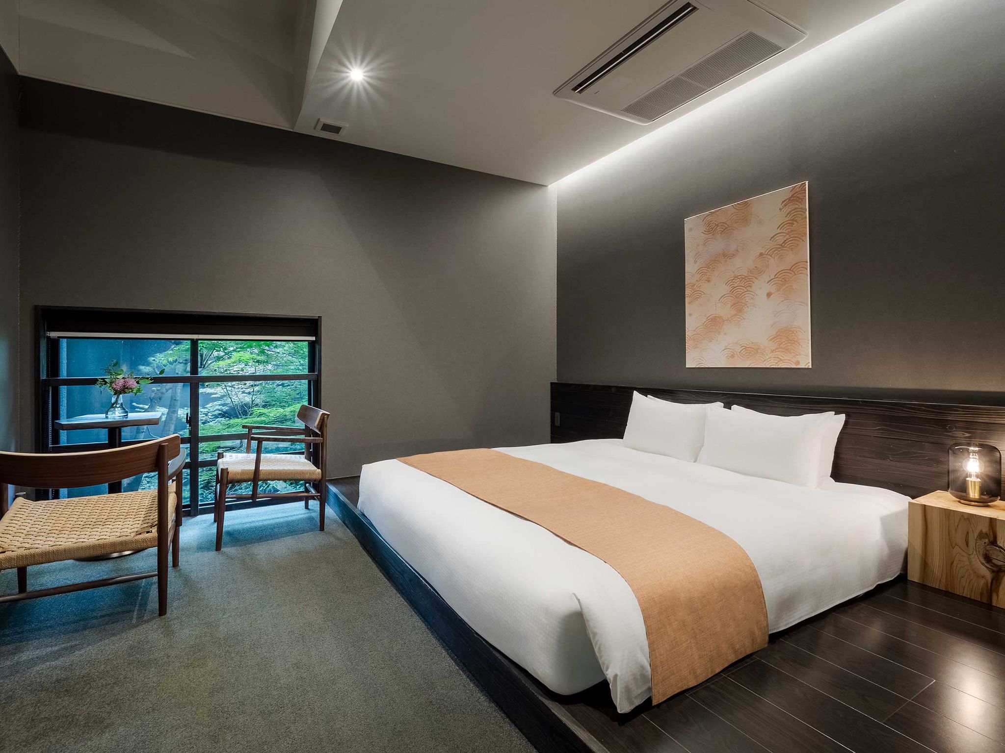 A hotel room with a large bed, a desk with a chair, a window with a view, and a wall-mounted artwork.