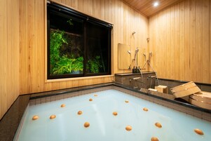 Interior de madera con una bañera grande que contiene bolas de madera, una ventana con vistas a la vegetación y una zona de ducha.