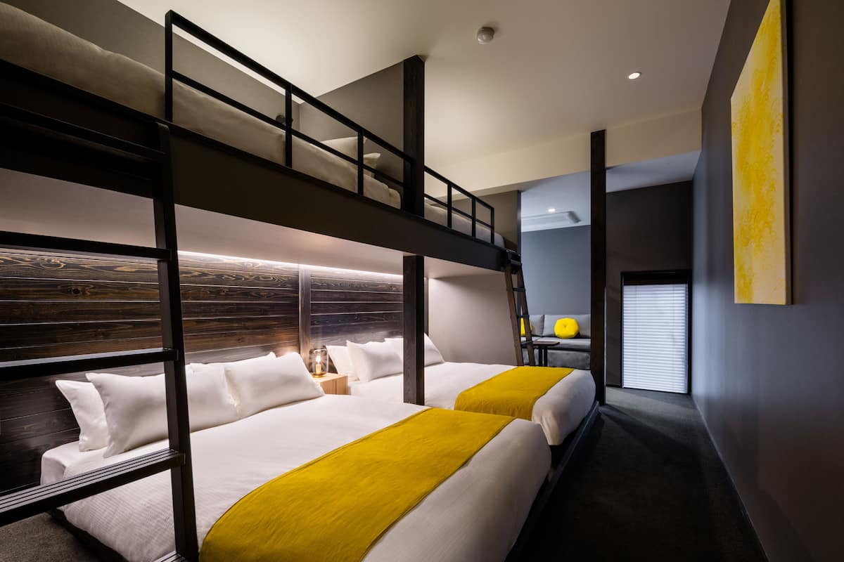 Een moderne hotelkamer met stapelbedden, een klein nachtkastje met lamp en een groot raam met zonwering.