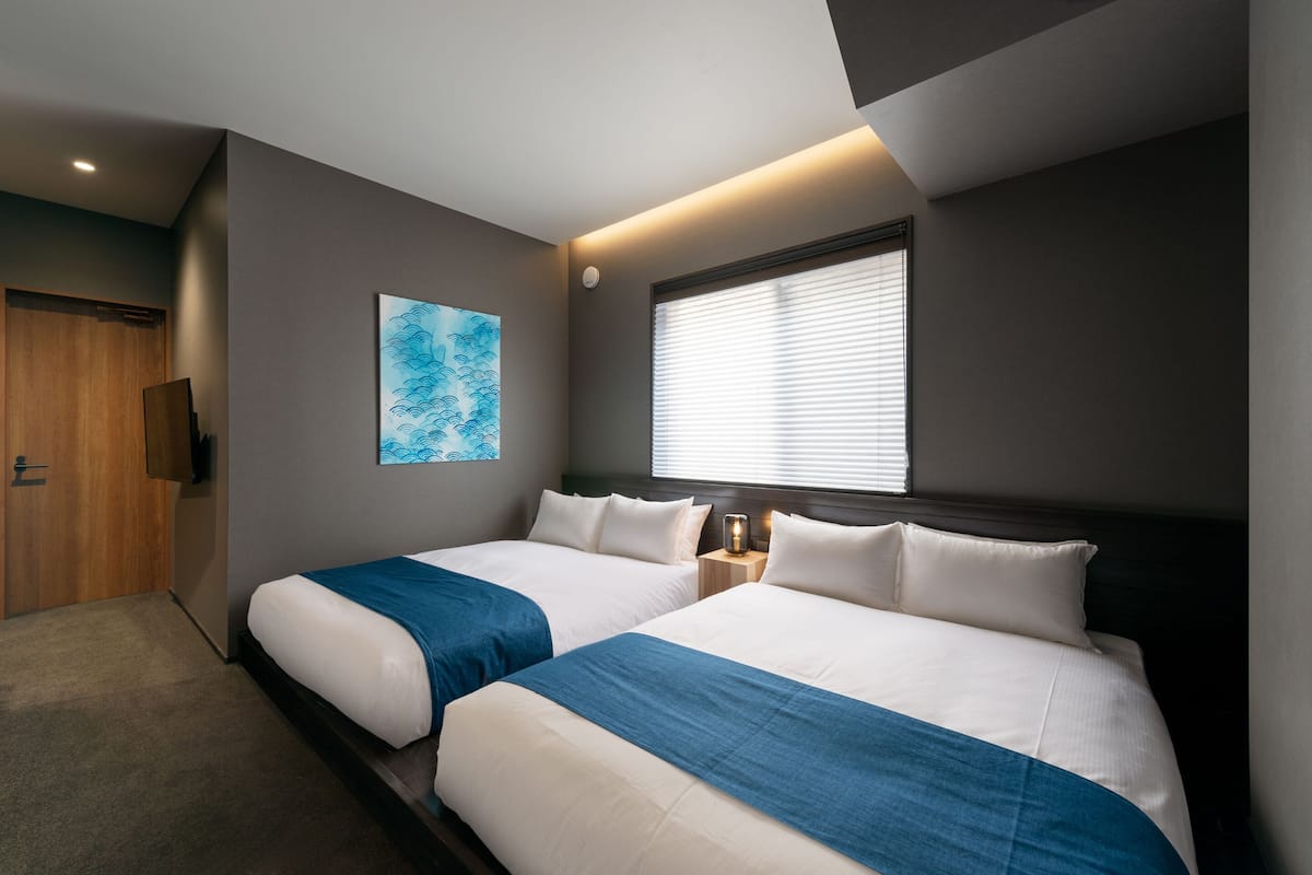Een hotelkamer met twee bedden, een houten garderobe, een schilderij en een raam met jaloezieën.
