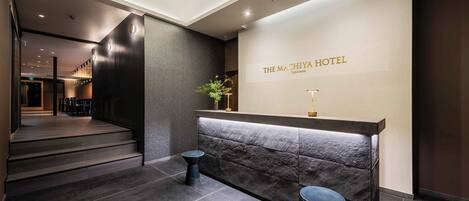 A hotel reception area with a modern design, featuring a reception desk, a staircase, and a wall-mounted sign.