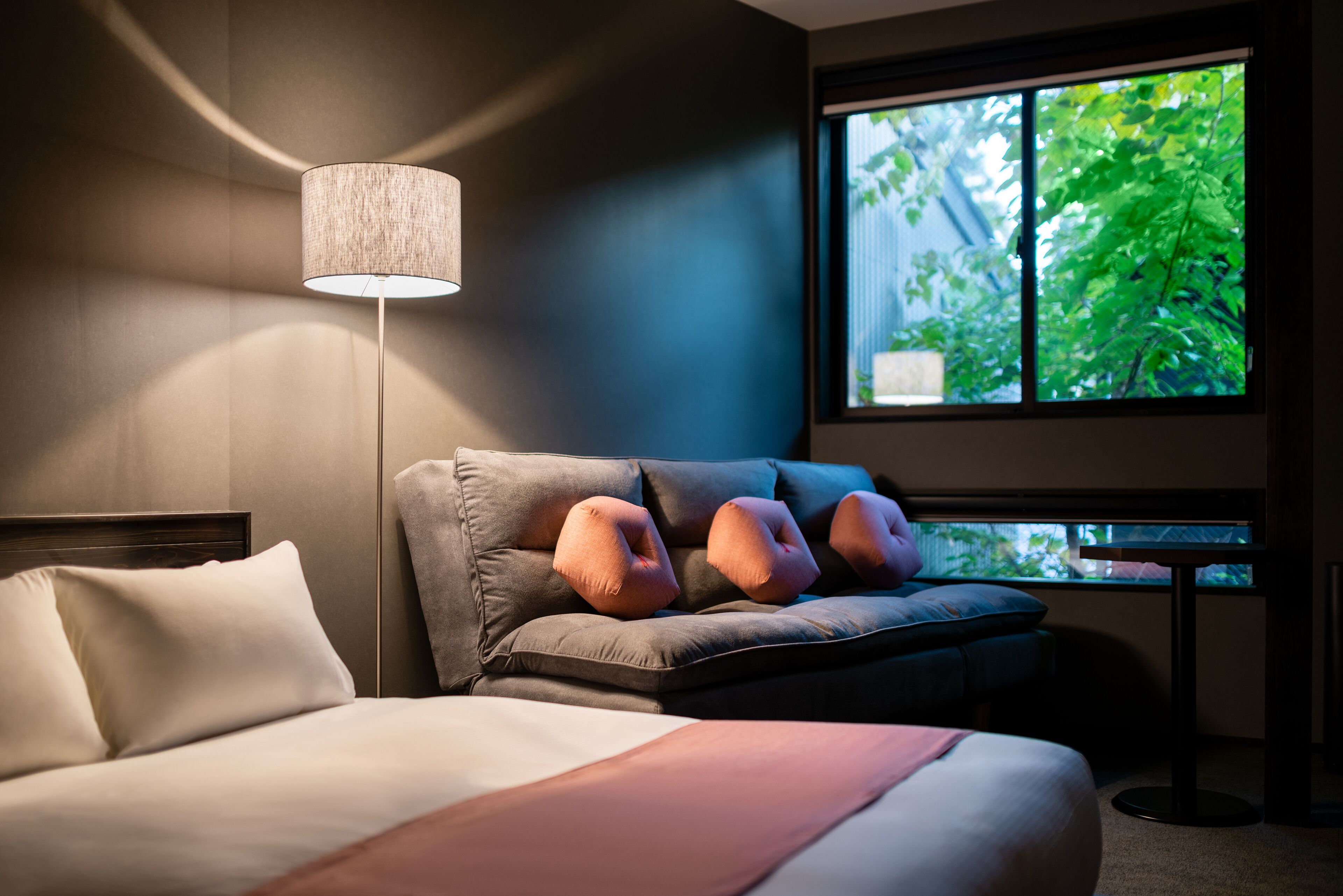 A modern hotel room with a bed, a sofa with decorative pillows, a standing lamp, and a window with a view of greenery.