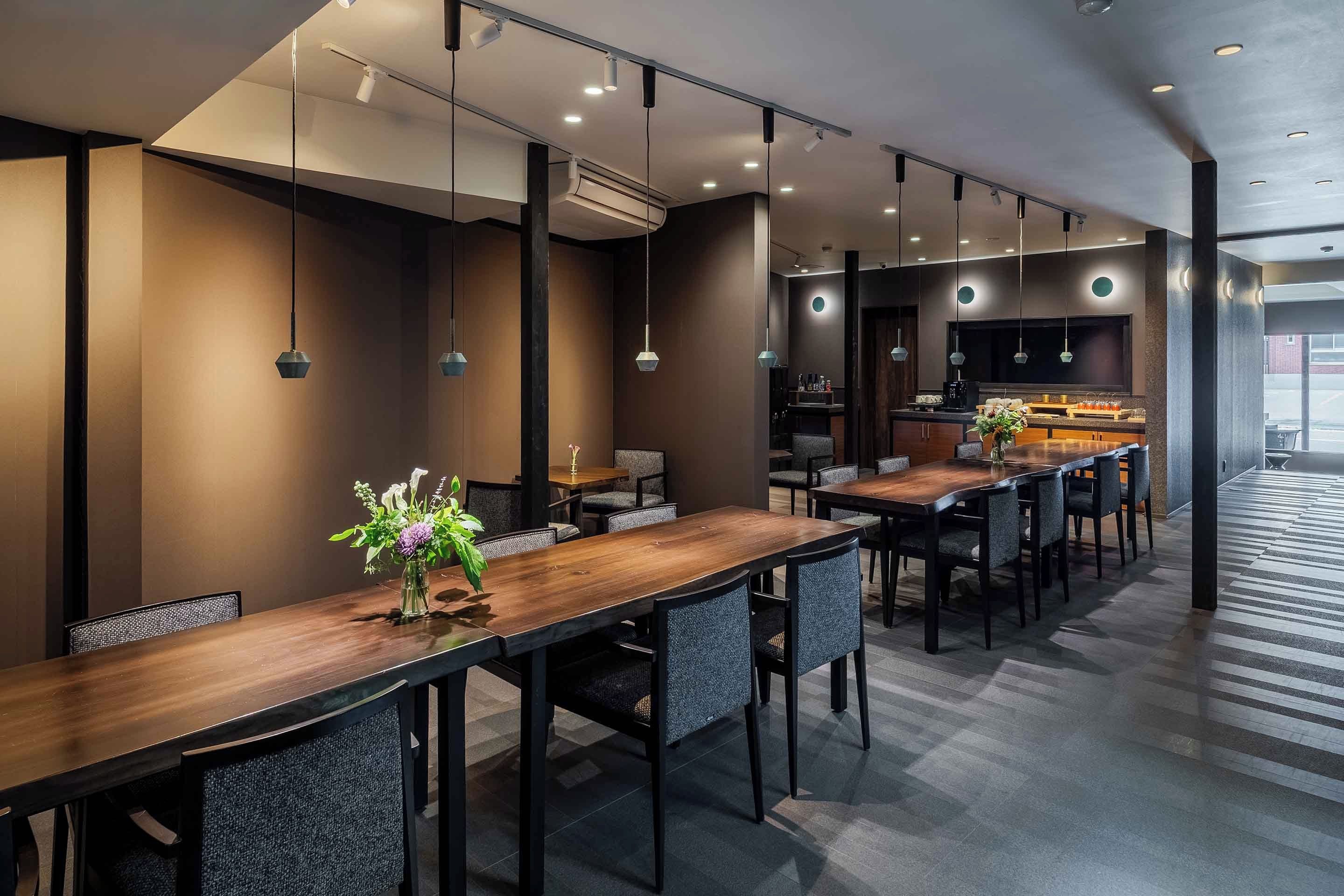 A modern dining area with long tables and chairs, recessed ceiling lighting, and a central island with a bar setup.
